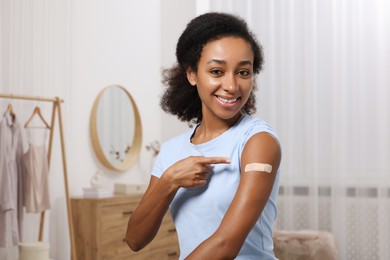 Photo of Happy young woman pointing at adhesive bandage after vaccination indoors