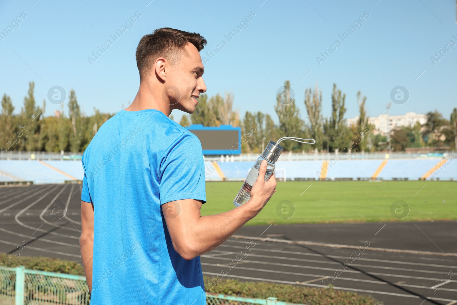 Photo of Young sporty man holding bottle of water at stadium on sunny day. Space for text