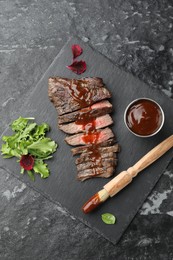 Photo of Pieces of delicious roasted beef meat with sauce and greens on black table, top view