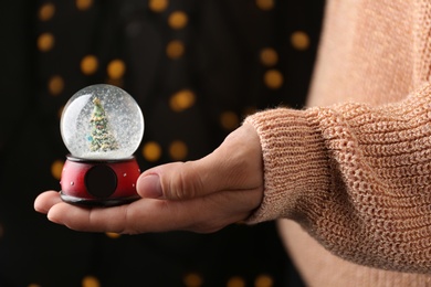 Photo of Woman holding snow globe with Christmas tree against festive lights, closeup