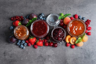 Jars with different jams and fresh fruits on grey table, flat lay