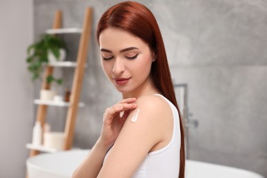 Photo of Beautiful young woman applying body cream onto shoulder in bathroom