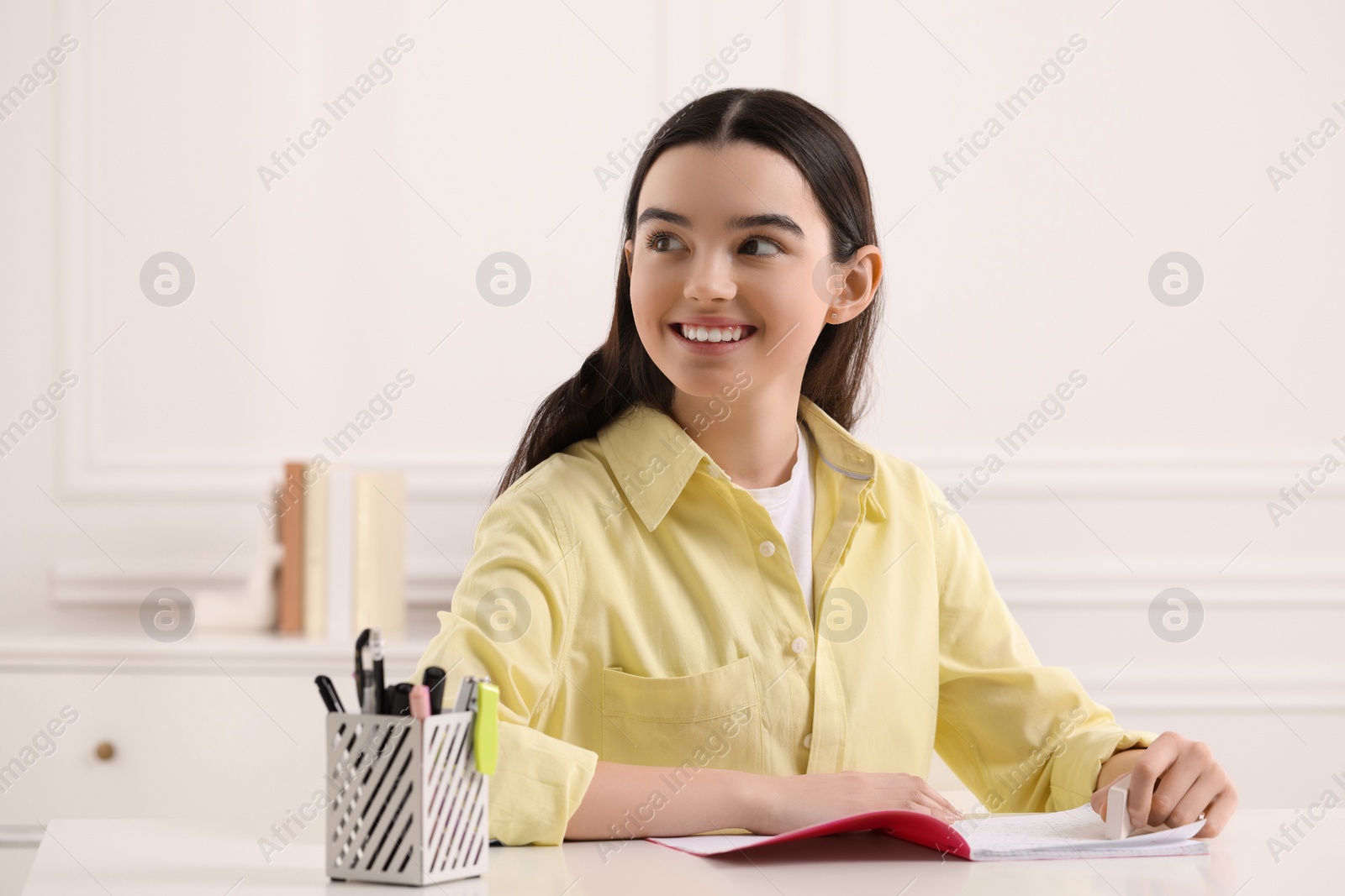 Photo of Teenage girl erasing mistake in her notebook at white desk indoors