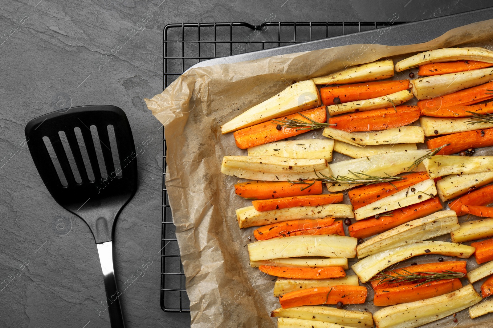 Photo of Tray with baked parsnips, carrots and spatula on black table, flat lay