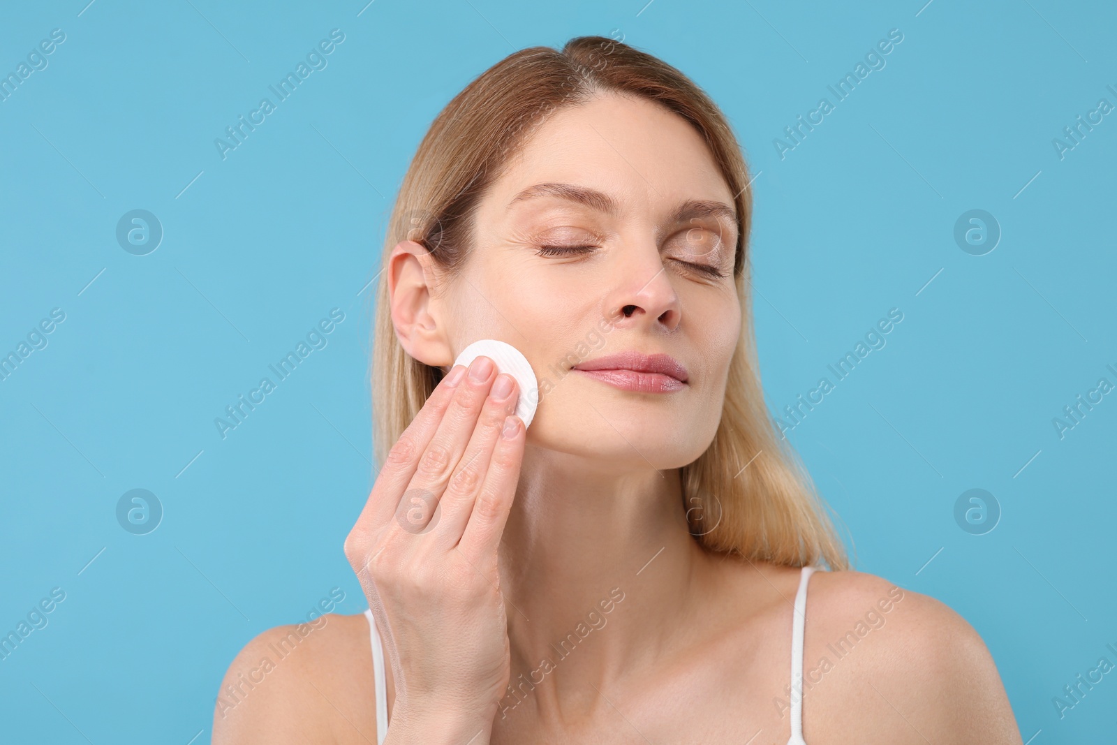 Photo of Beautiful woman removing makeup with cotton pad on light blue background