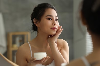 Happy woman applying face cream near mirror at home