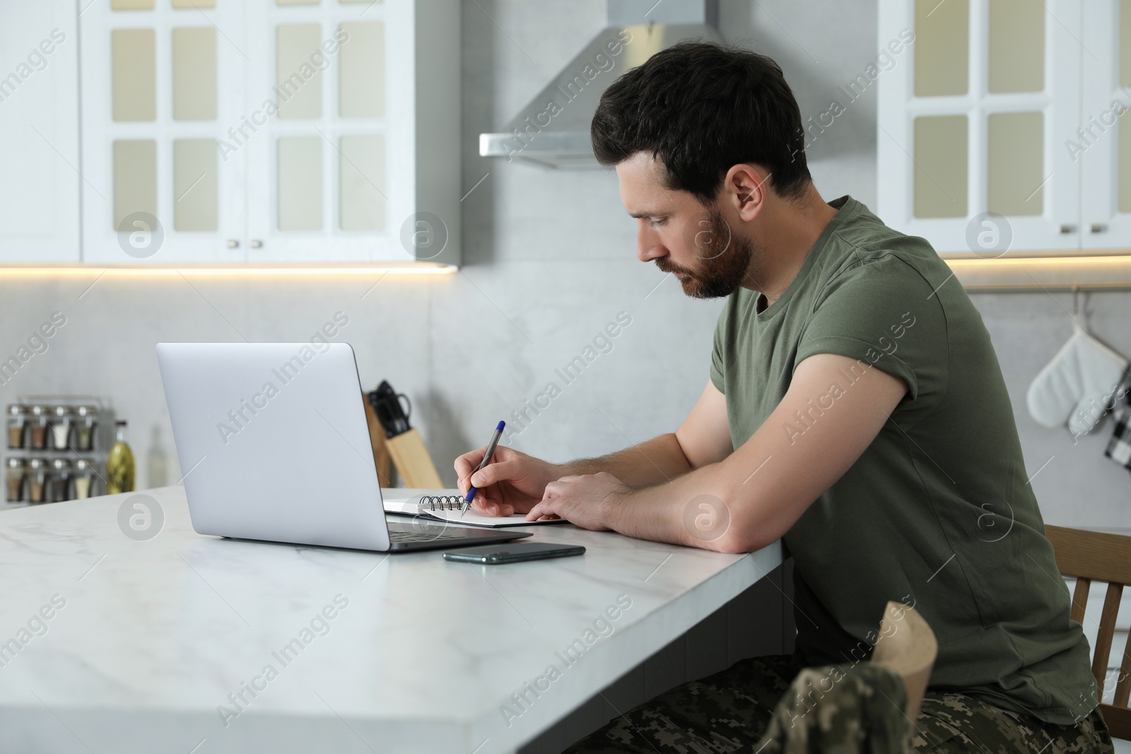 Photo of Soldier taking notes while working with laptop at white marble table in kitchen. Military service