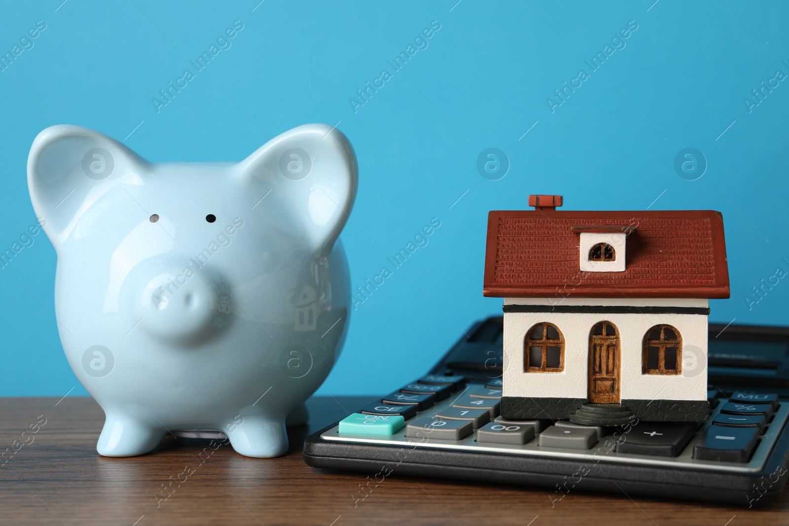 Photo of House model, piggy bank and calculator on wooden table against light blue background