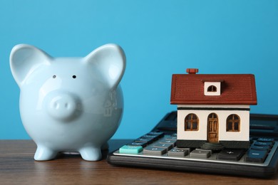 House model, piggy bank and calculator on wooden table against light blue background