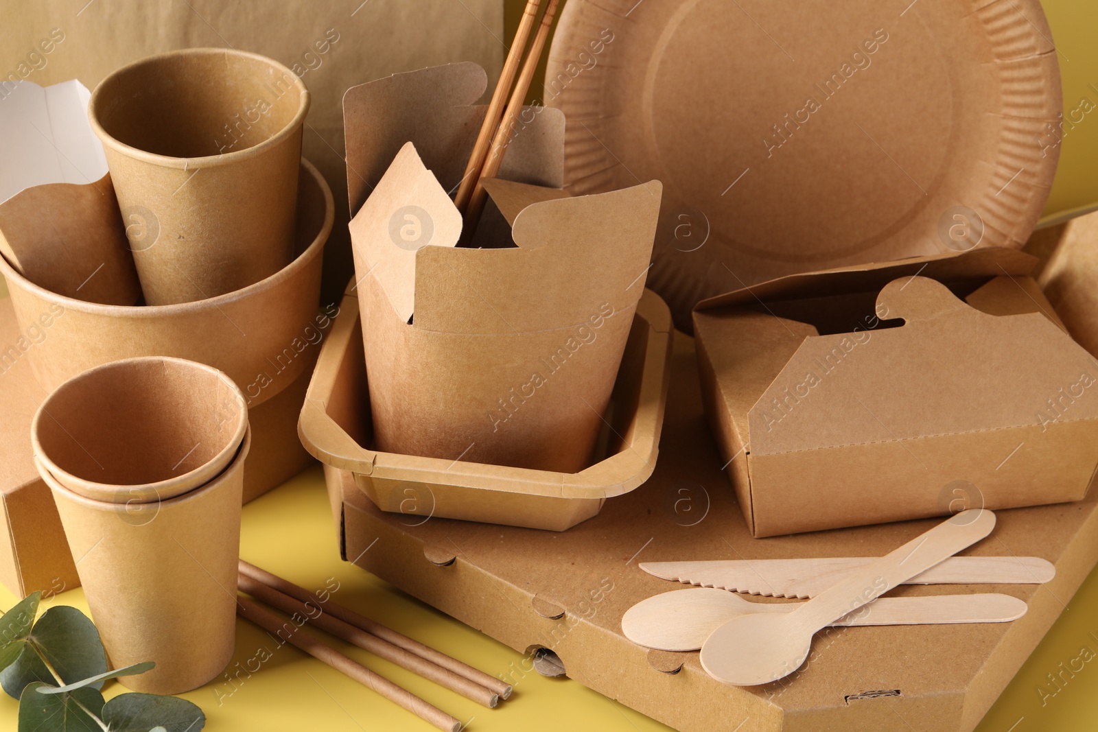 Photo of Eco friendly food packaging. Paper containers, tableware and eucalyptus leaves on pale yellow background, closeup