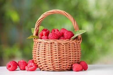 Photo of Wicker basket with tasty ripe raspberries and leaves on white table against blurred green background