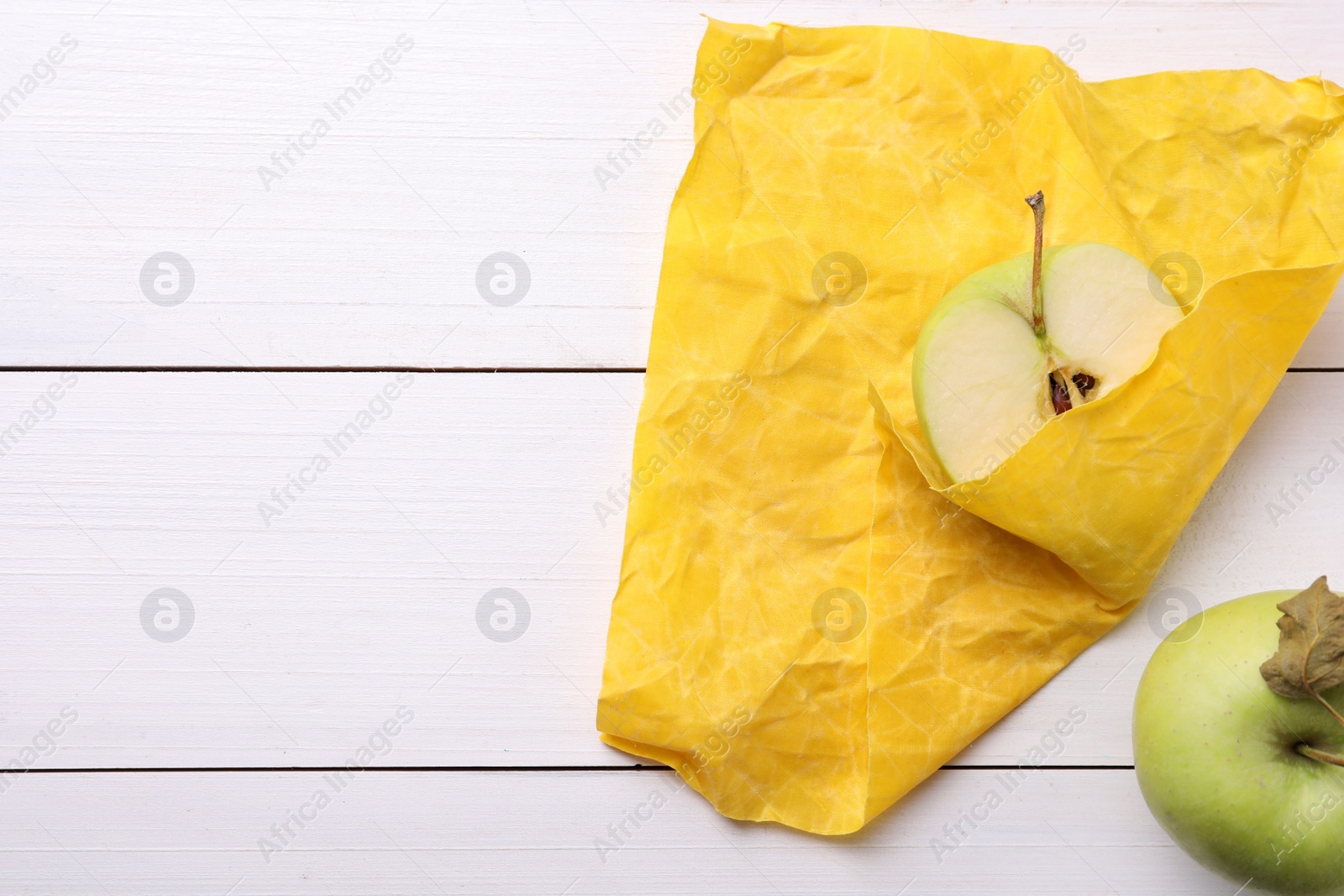 Photo of Half of apple in yellow beeswax food wrap on white wooden table, top view. Space for text