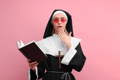 Photo of Surprised woman in nun habit and sunglasses holding Bible against pink background
