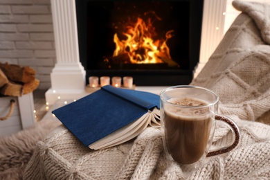 Cup of coffee and book on plaid near fireplace indoors. Cozy atmosphere