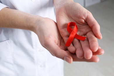 Photo of Doctor holding red awareness ribbon indoors, closeup. World AIDS disease day