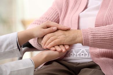 Photo of Nurse holding hands of elderly woman against blurred background, closeup. Assisting senior generation