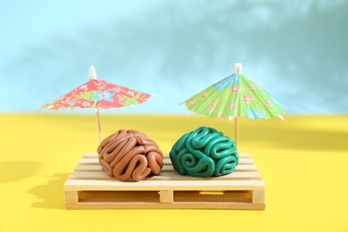 Photo of Brains made of plasticine on mini wooden sunbed under umbrellas against color background