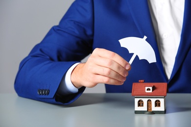 Photo of Man covering house model with umbrella cutout at table, closeup. Home insurance