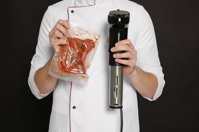 Chef holding sous vide cooker and meat in vacuum pack on black background, closeup