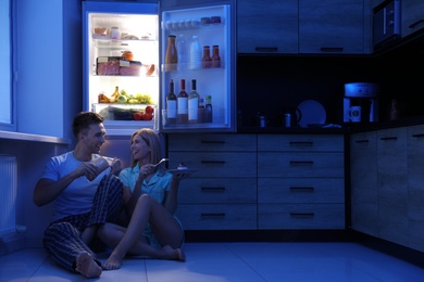 Photo of Happy couple eating near refrigerator in kitchen at night