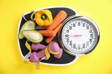 Photo of Scales, healthy food, dumbbells and measuring tape on yellow background, top view