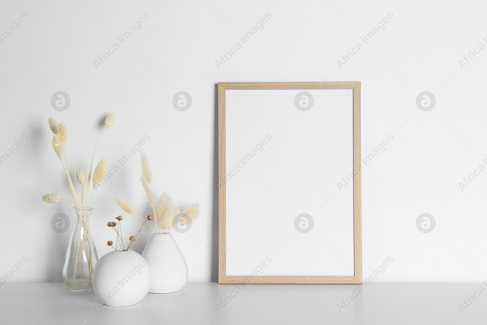 Photo of Empty photo frame and vases with dry decorative spikes on white table. Mockup for design