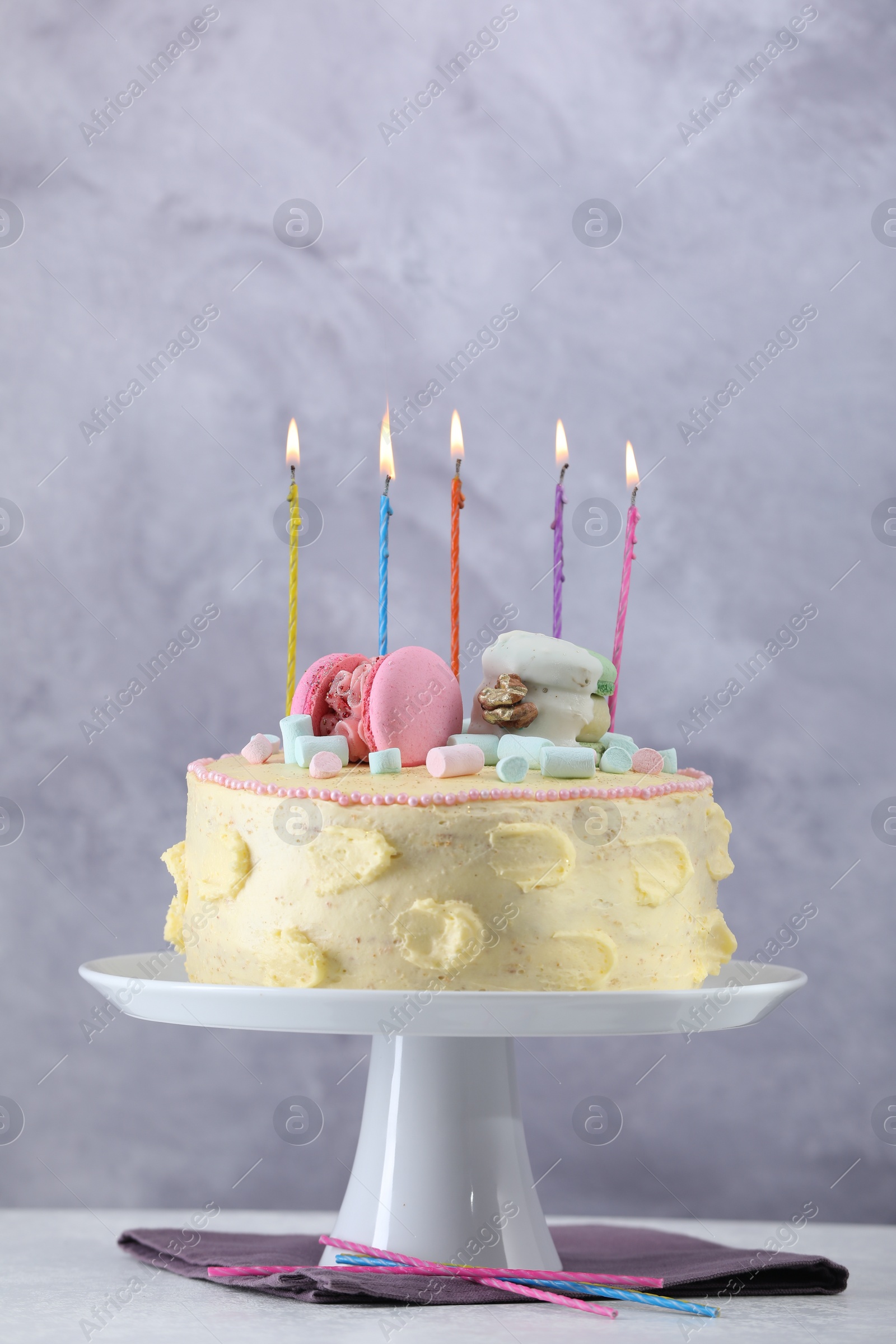 Photo of Delicious cake decorated with macarons, marshmallows and burning candles served on white table against grey background