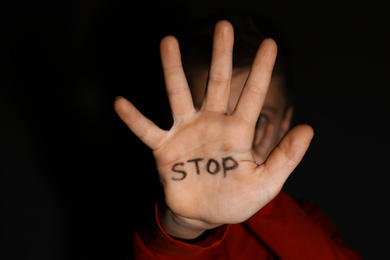 Abused little boy showing palm with word STOP against black background, focus on hand. Domestic violence concept