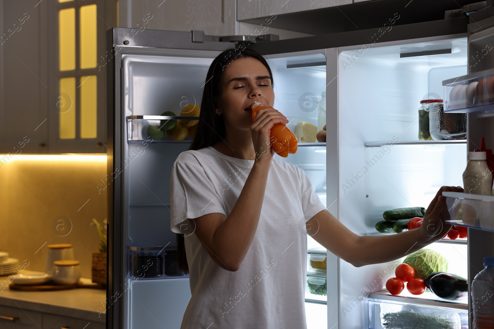 Photo of Young woman drinking juice near modern refrigerator in kitchen at night