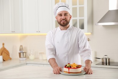 Photo of Happy professional confectioner with delicious cake at table in kitchen