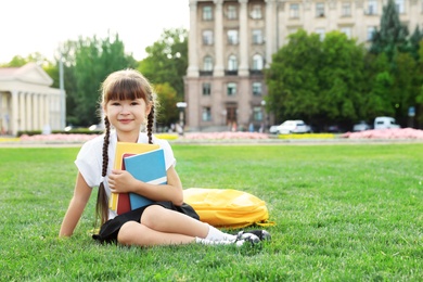 Cute girl with school stationery sitting on green lawn outdoors