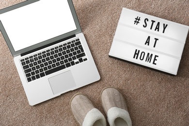 Photo of Laptop, slippers and lightbox with hashtag STAY AT HOME on soft carpet, flat lay. Message to promote self-isolation during COVID‑19 pandemic