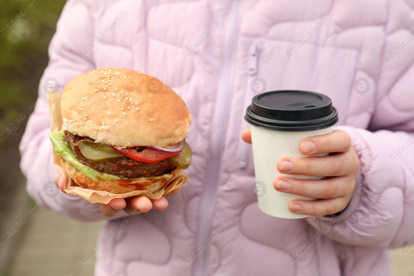 Photo of Little girl holding fresh delicious burger and cup of coffee outdoors, closeup. Street food