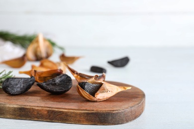 Photo of Wooden board with black garlic on white table. Space for text