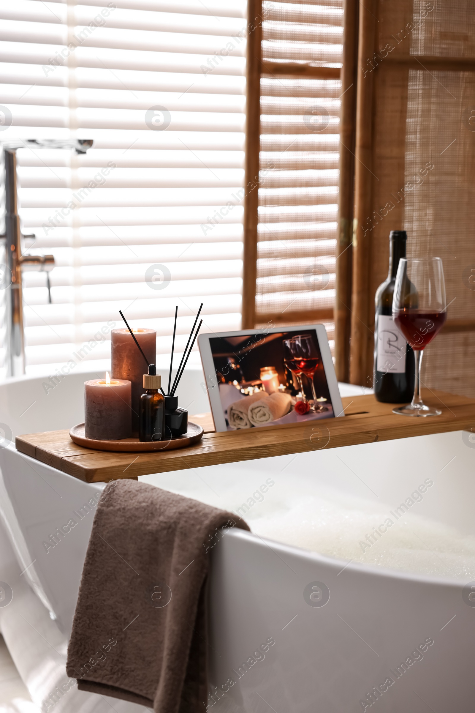 Photo of Wooden tray with tablet, wine and burning candles on bathtub in bathroom