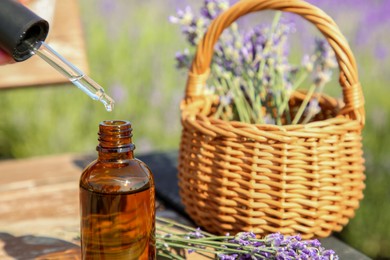 Dripping essential oil from pipette into bottle near lavender on wooden table outdoors, closeup. Space for text