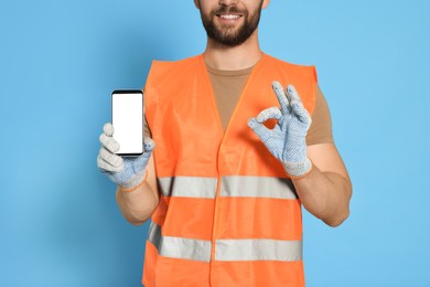 Man in reflective uniform showing smartphone and OK gesture on light blue background, closeup