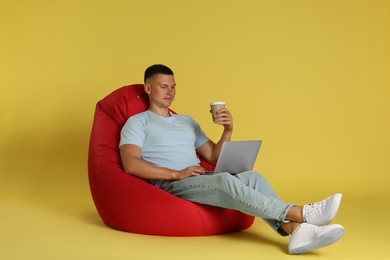 Photo of Happy man with laptop drinking coffee on red bean bag chair against yellow background