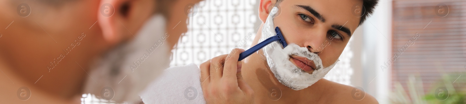 Image of Handsome young man shaving with razor near mirror in bathroom. Banner design