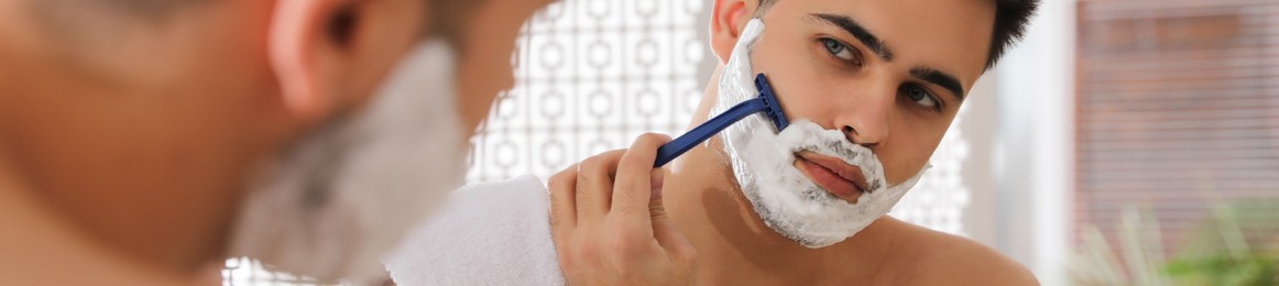 Image of Handsome young man shaving with razor near mirror in bathroom. Banner design