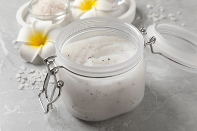 Photo of Body scrub in glass jar and plumeria flowers on grey marble table, closeup