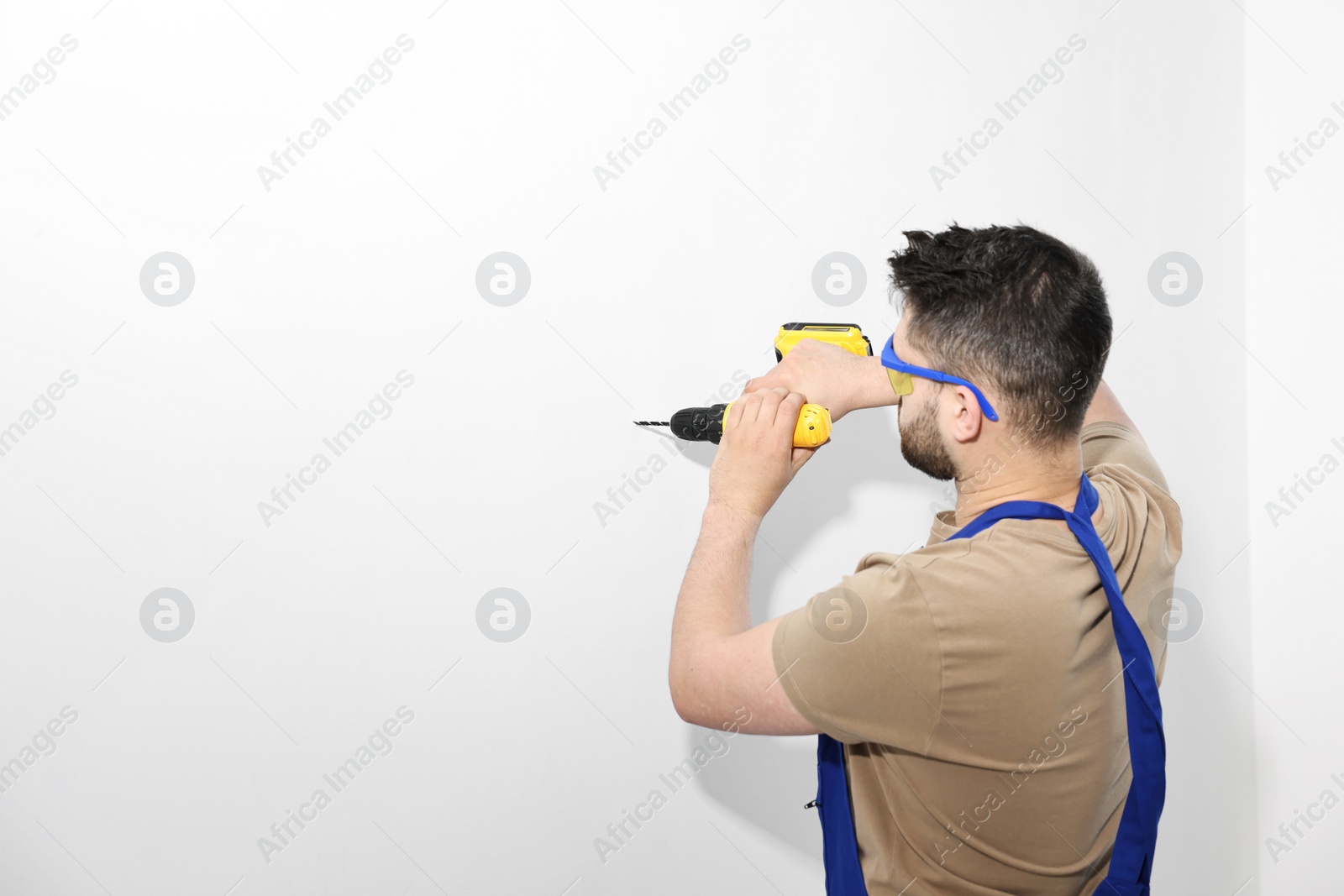 Photo of Young worker in uniform using electric drill indoors. Space for text