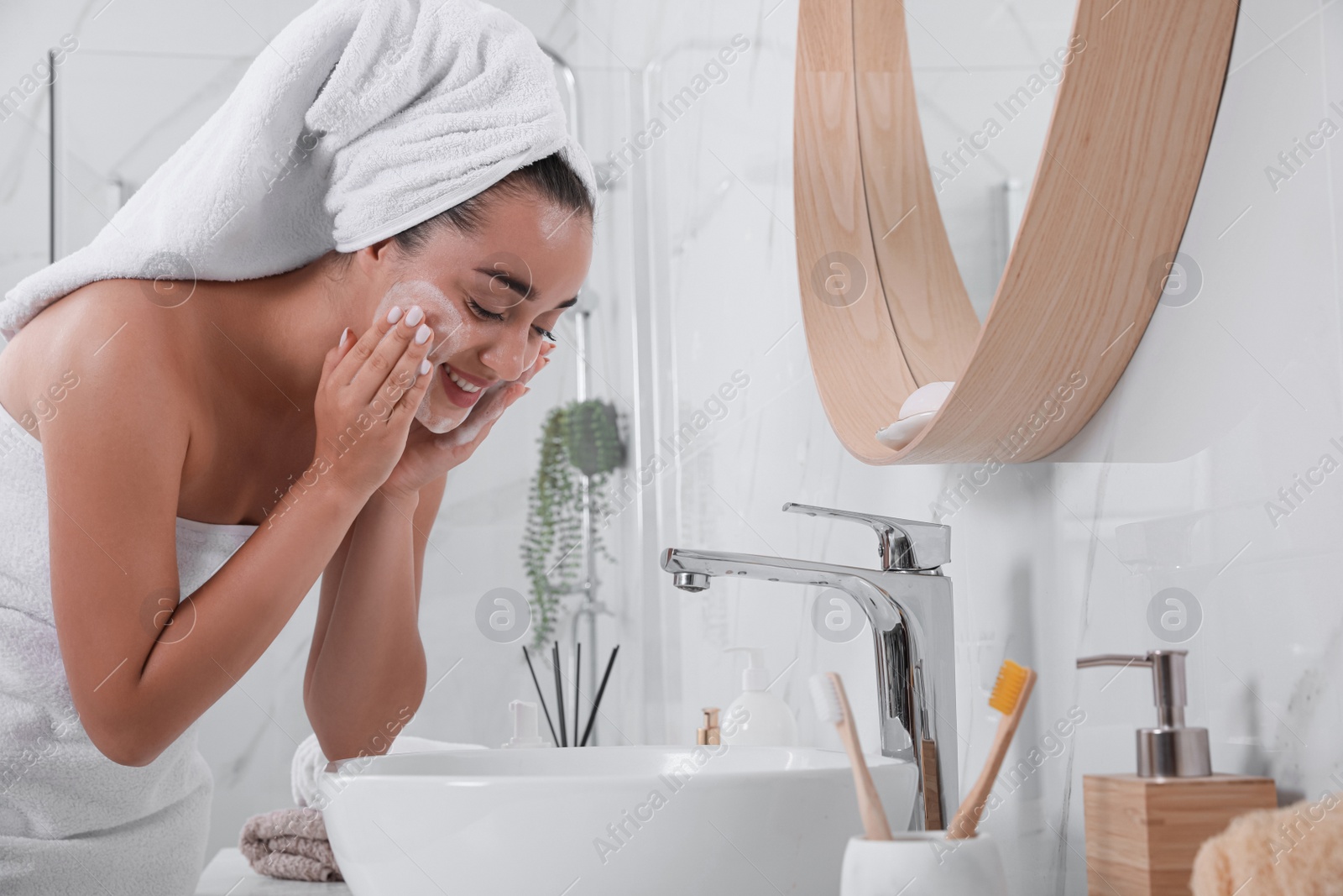 Photo of Beautiful young woman applying cleansing foam onto face near mirror in bathroom. Skin care cosmetic