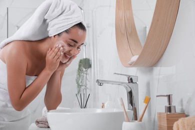 Beautiful young woman applying cleansing foam onto face near mirror in bathroom. Skin care cosmetic