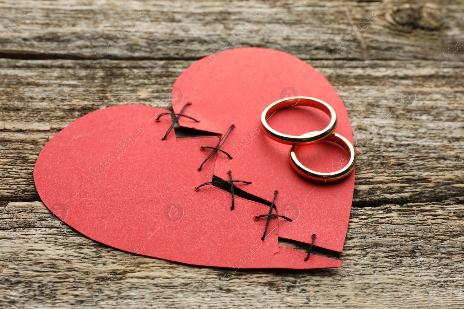 Photo of Broken heart. Torn red paper heart sewed with thread and wedding rings on wooden table, closeup