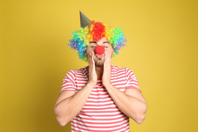 Photo of Emotional young man with party hat and clown wig on yellow background. April fool's day