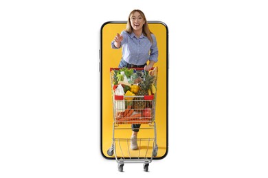 Image of Grocery shopping via internet. Happy woman with shopping cart full of products pointing at something while walking out of huge smartphone on white background