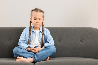 Little girl meditating on soft sofa indoors. Space for text