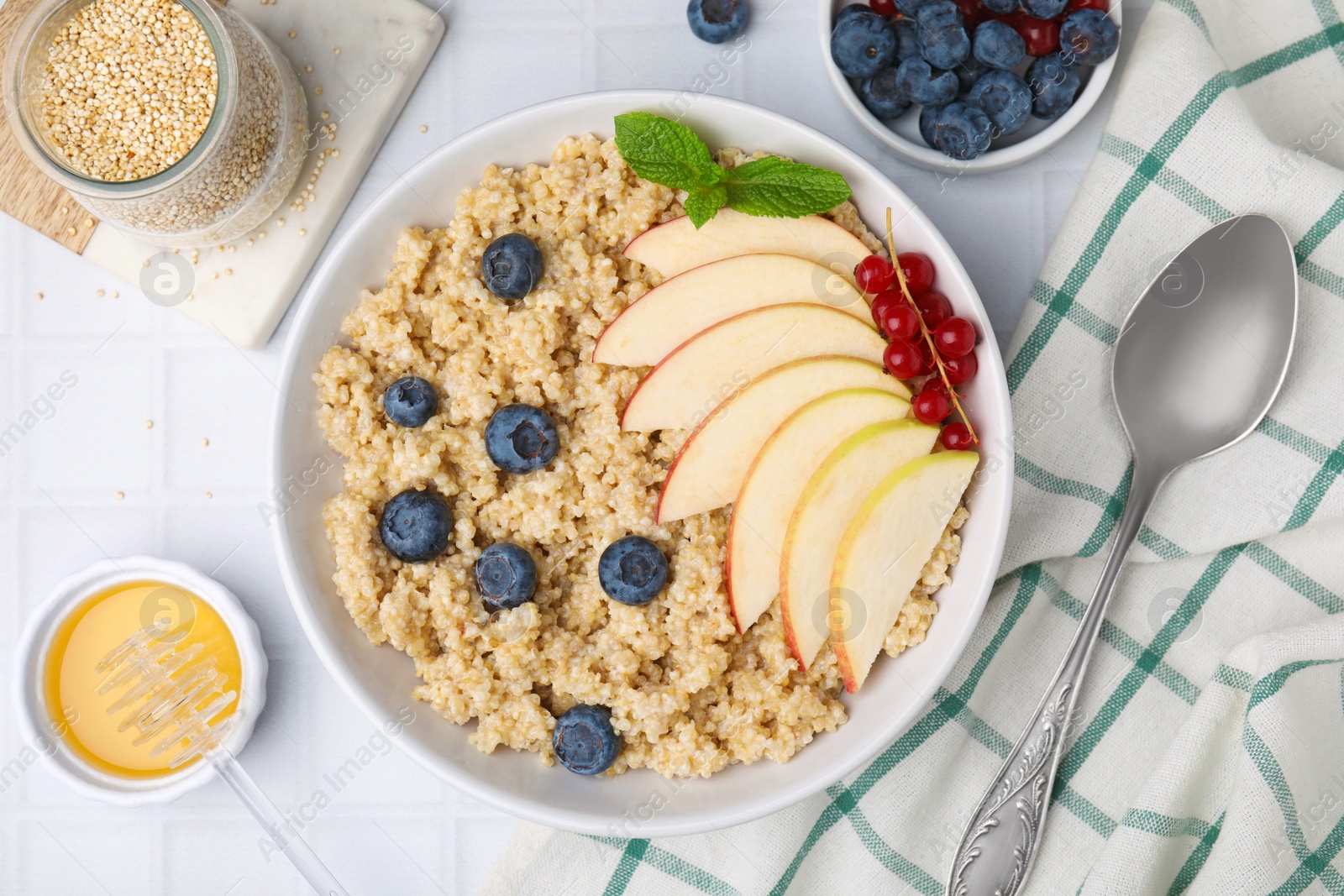 Photo of Bowl of delicious cooked quinoa with apples, blueberries and cranberries on white tiled table, flat lay