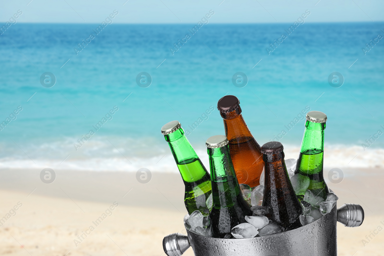 Image of Bottles of beer with ice cubes in metal bucket against blurred ocean and sandy beach